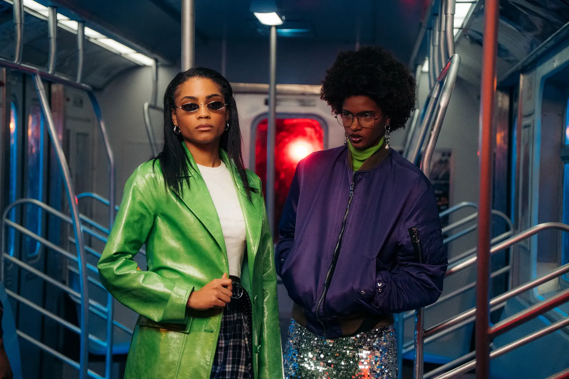   Two models pose side by side in a subway set, one wearing a green coat and the other a purple jacket with a silver skirt.