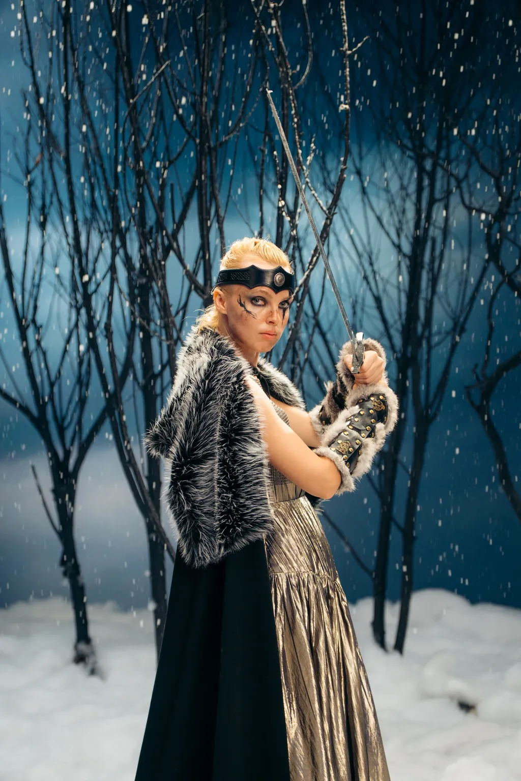 A model dressed as a warrior holds a raised sword, wearing a fur-trimmed cape in a snowy forest set.