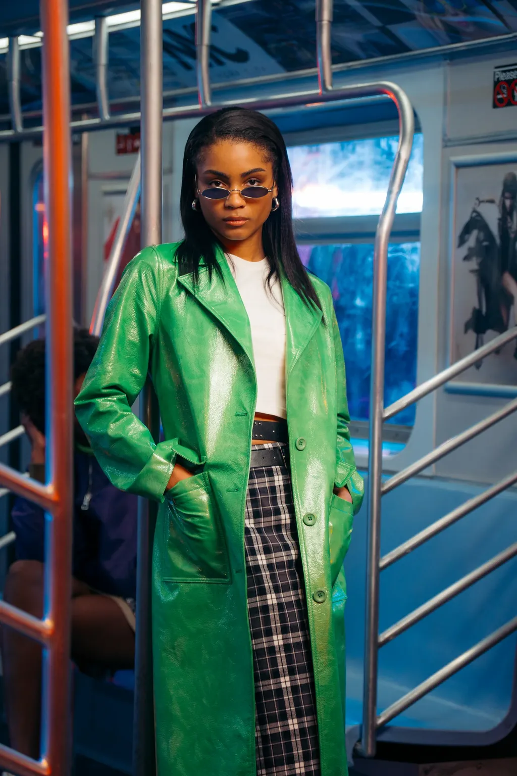 A model in a glossy green coat, plaid skirt and narrow sunglasses poses with her hands in her pockets in a subway set.