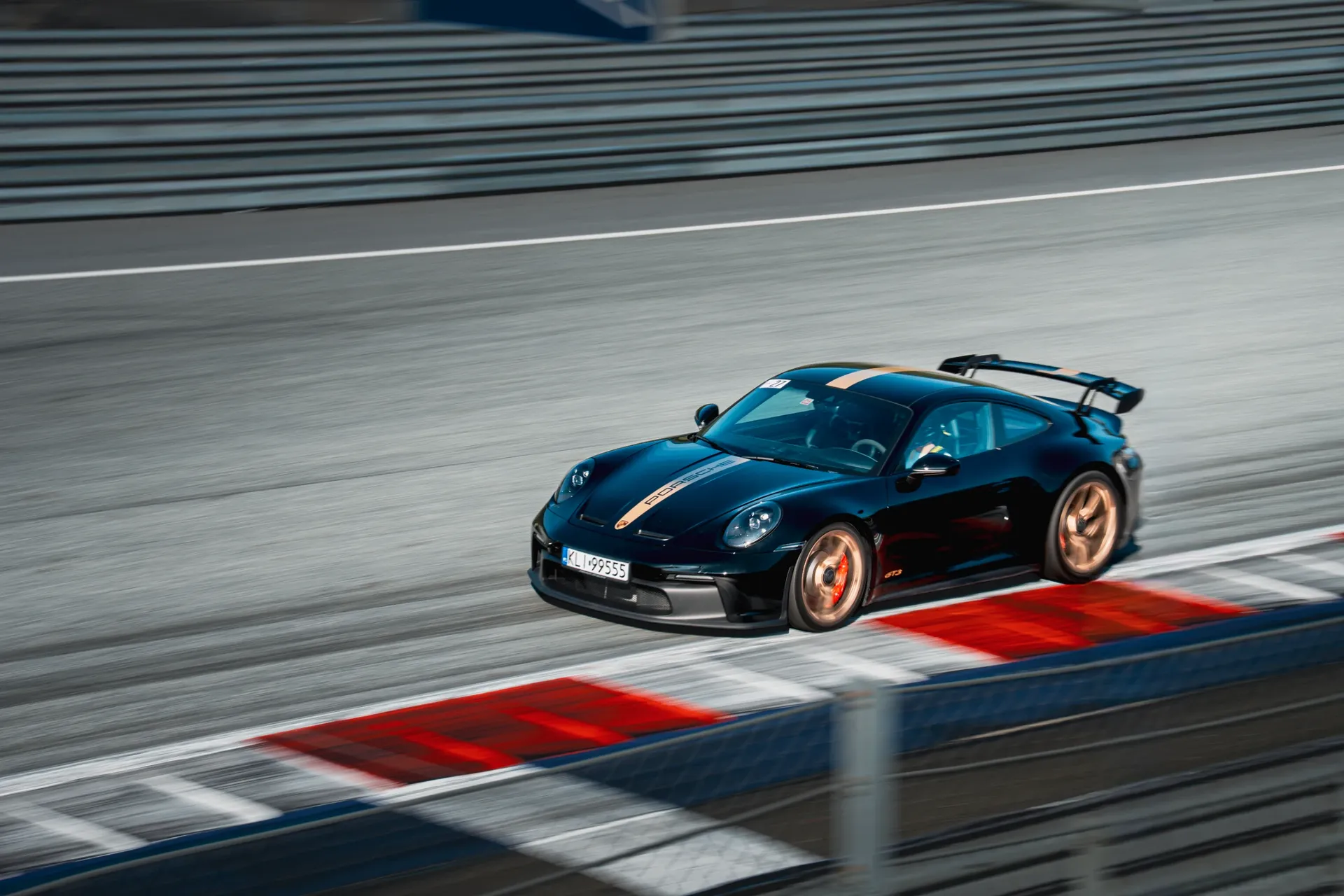 Black Porsche 911 GT3 with gold wheels and a gold stripe driving beside the red and white curbing at the Red Bull Ring.