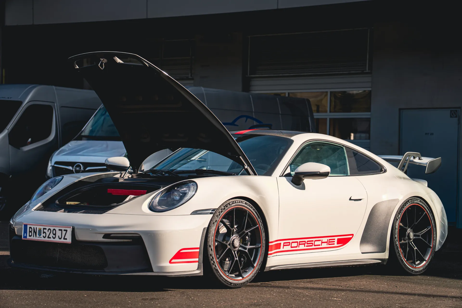 White Porsche 911 with red side stripes and its front luggage compartment open outside the Red Bull Ring garages.
