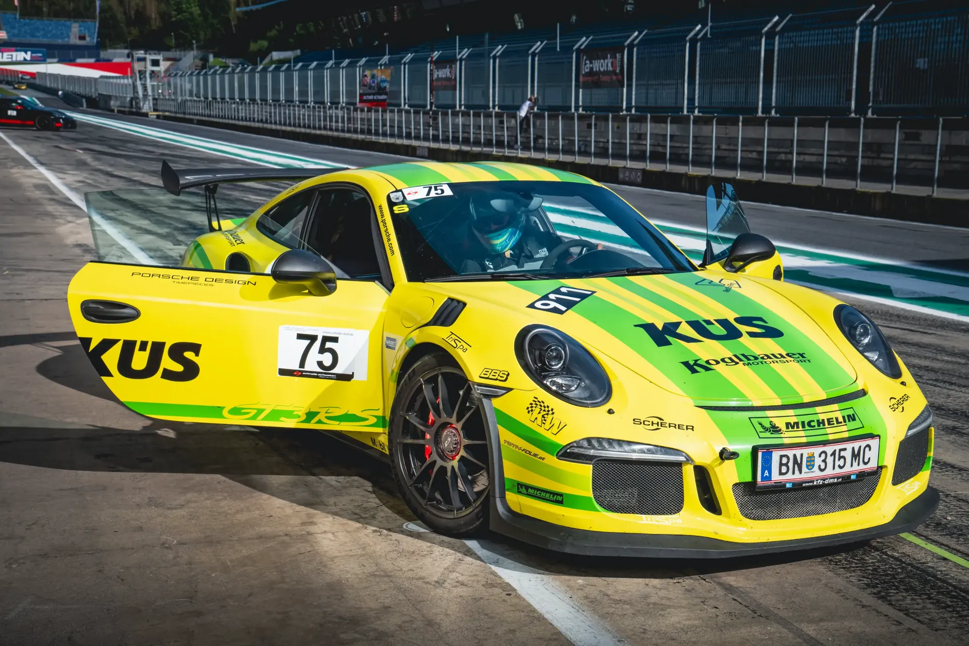 Yellow and green Porsche 911 GT3 RS with both doors open and a helmeted driver inside at the Red Bull Ring.