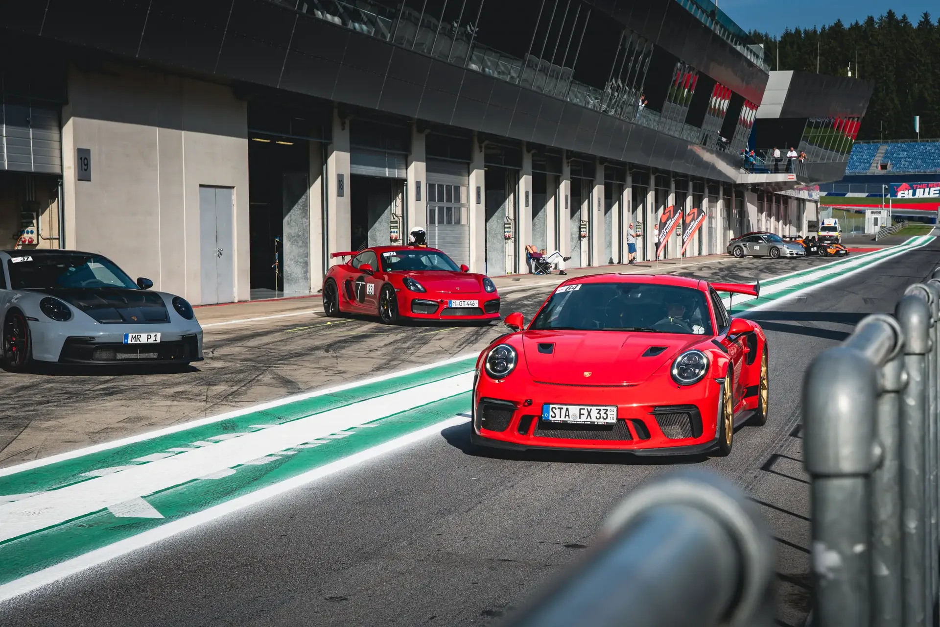 Red Porsche 911 GT3 RS driving through the Red Bull Ring pit lane, with other Porsches parked outside the garages.