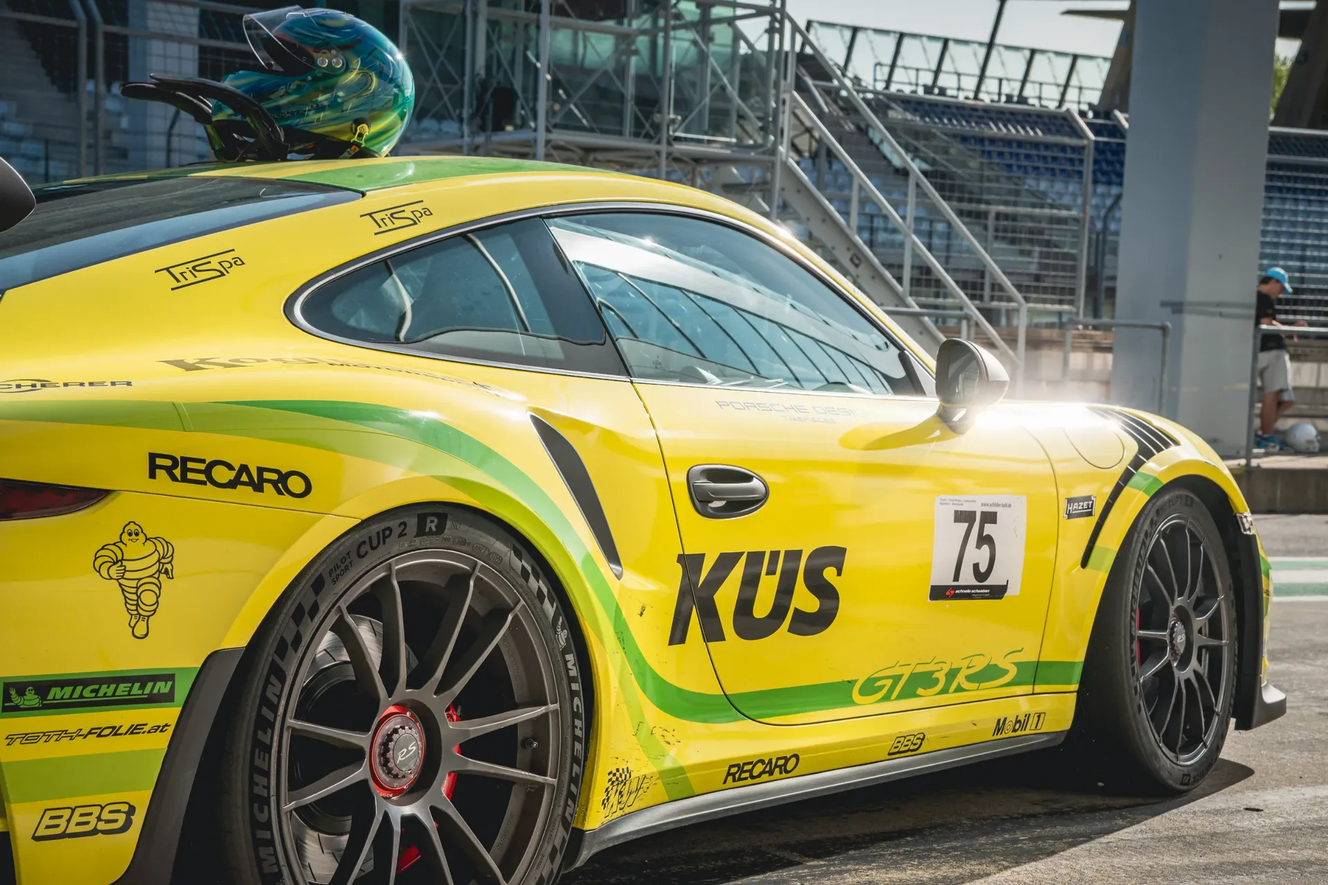 Side detail of a yellow and green Porsche 911 GT3 RS with racing decals and a helmet resting on the roof.