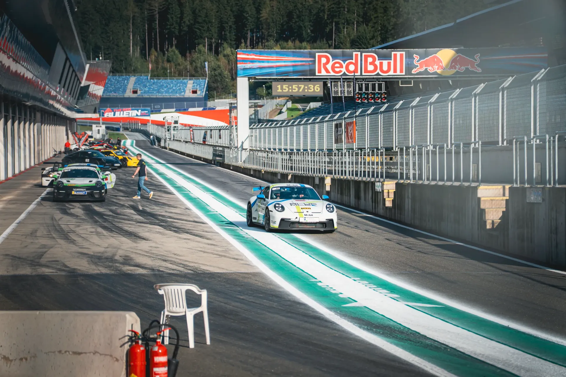 White Porsche with blue accents driving along the Red Bull Ring pit lane, with a row of sports cars beside the garages.