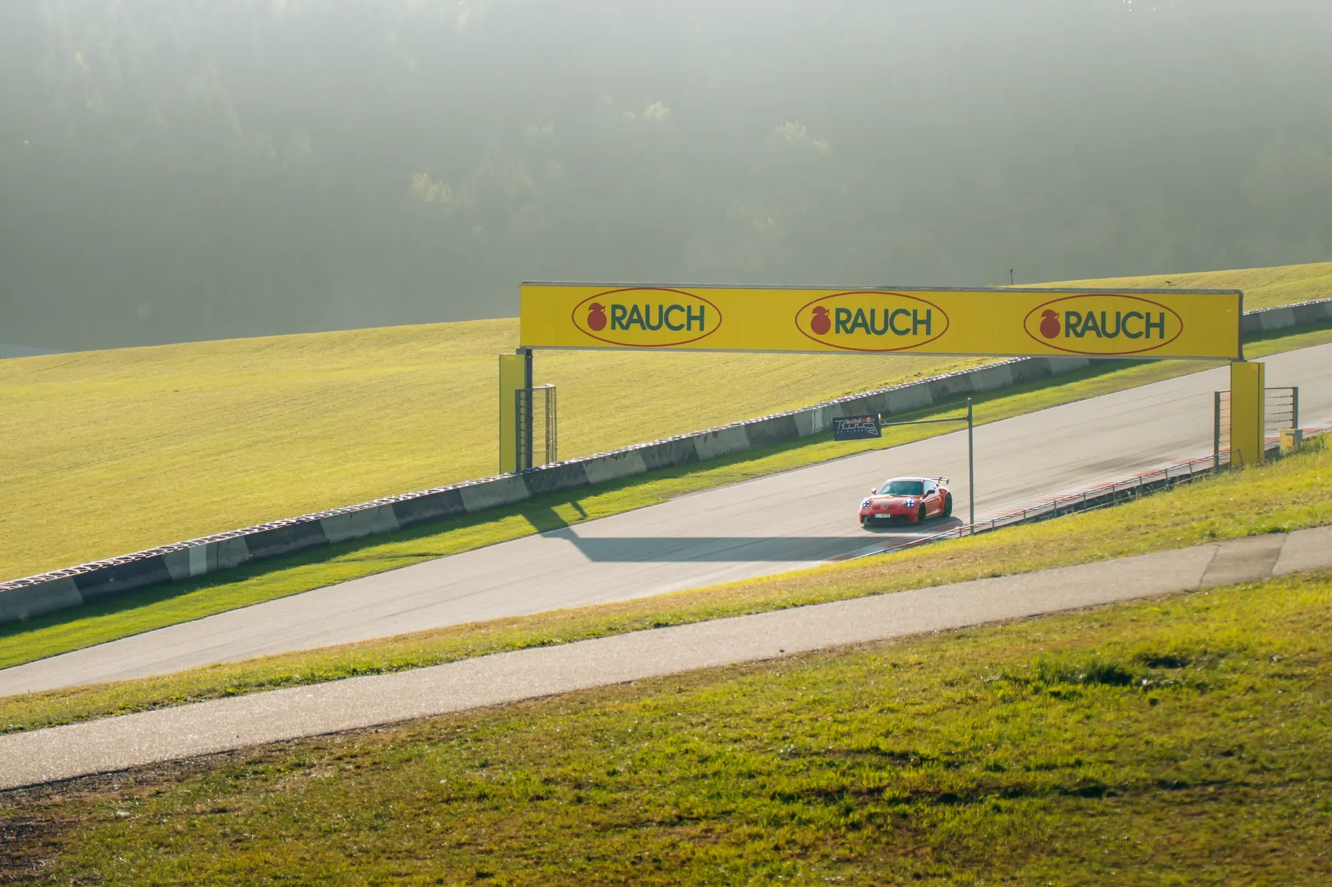 Red Porsche driving beneath a yellow track banner at the Red Bull Ring, surrounded by sunlit grass and wooded hills.