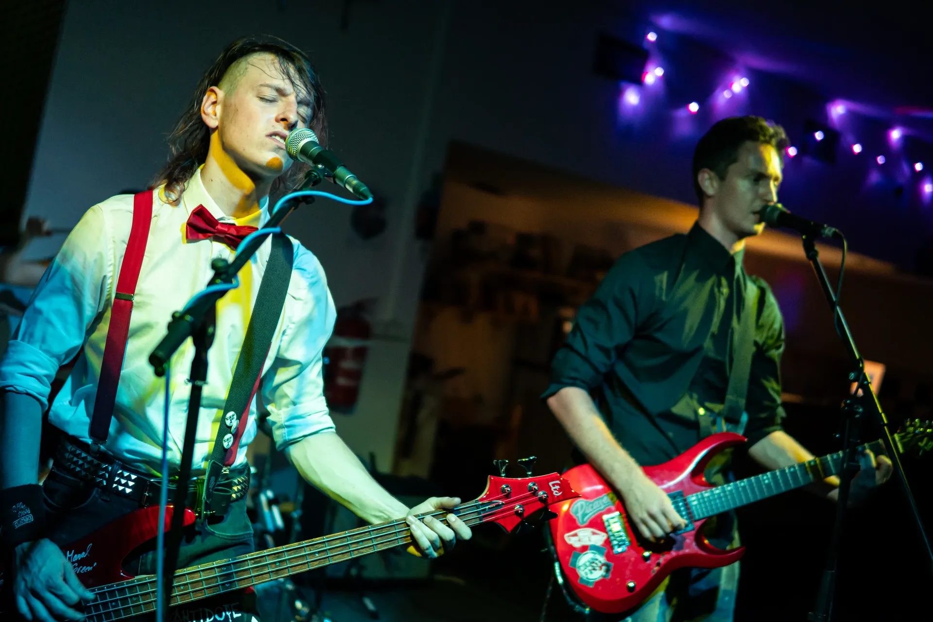 Emergency Brake’s bassist and guitarist sing into microphones while playing red instruments under colorful stage lights.