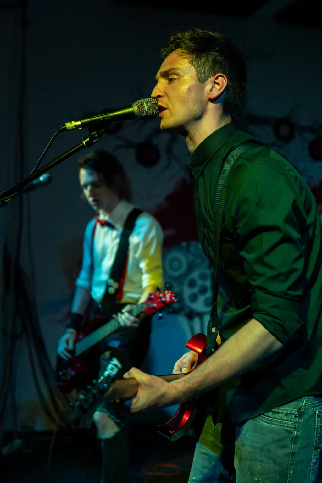 Emergency Brake’s guitarist sings into a microphone under green stage lighting, with the bassist in the background.