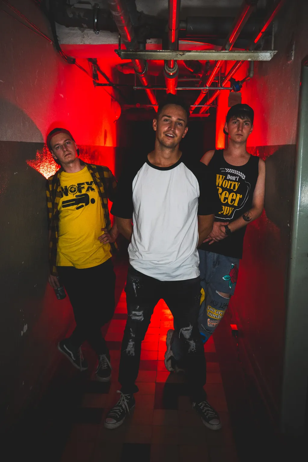 The three members of Emergency Brake pose in a narrow corridor with exposed ceiling pipes and red backlighting.
