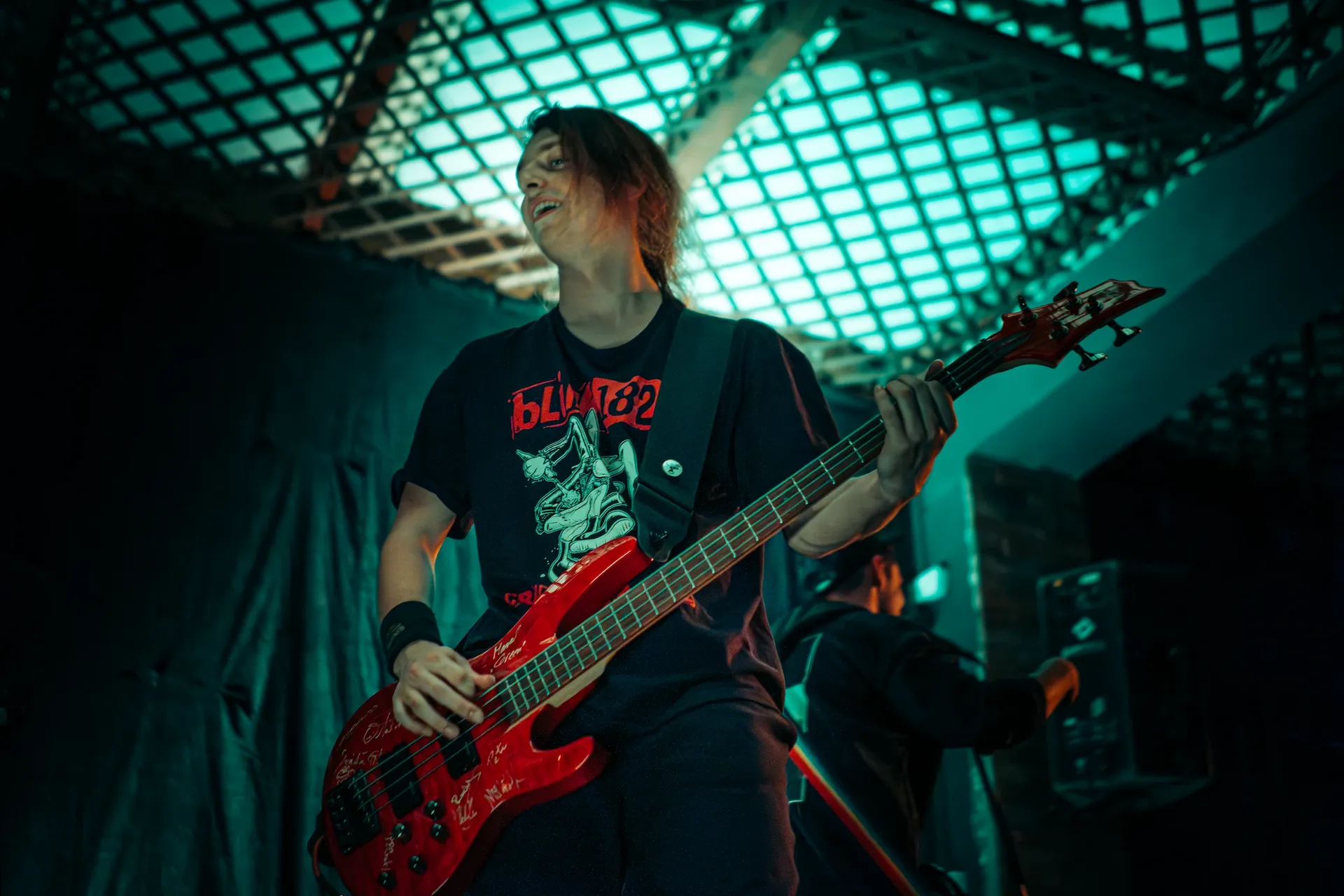 Emergency Brake’s bassist plays a red bass guitar under turquoise lighting while wearing a blink-182 T-shirt.